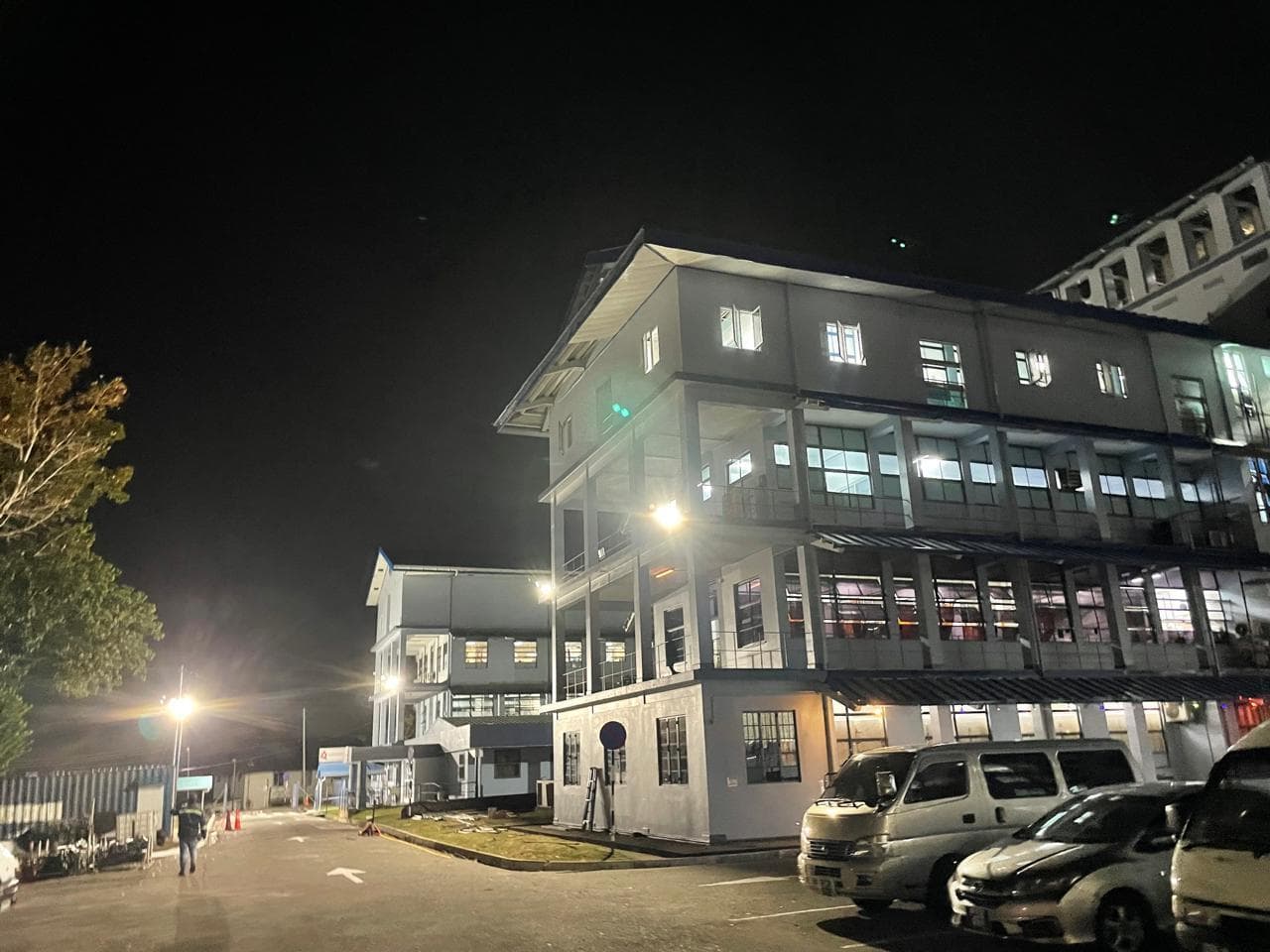 Illuminated multi-story building at night with parked cars and vans in the foreground.