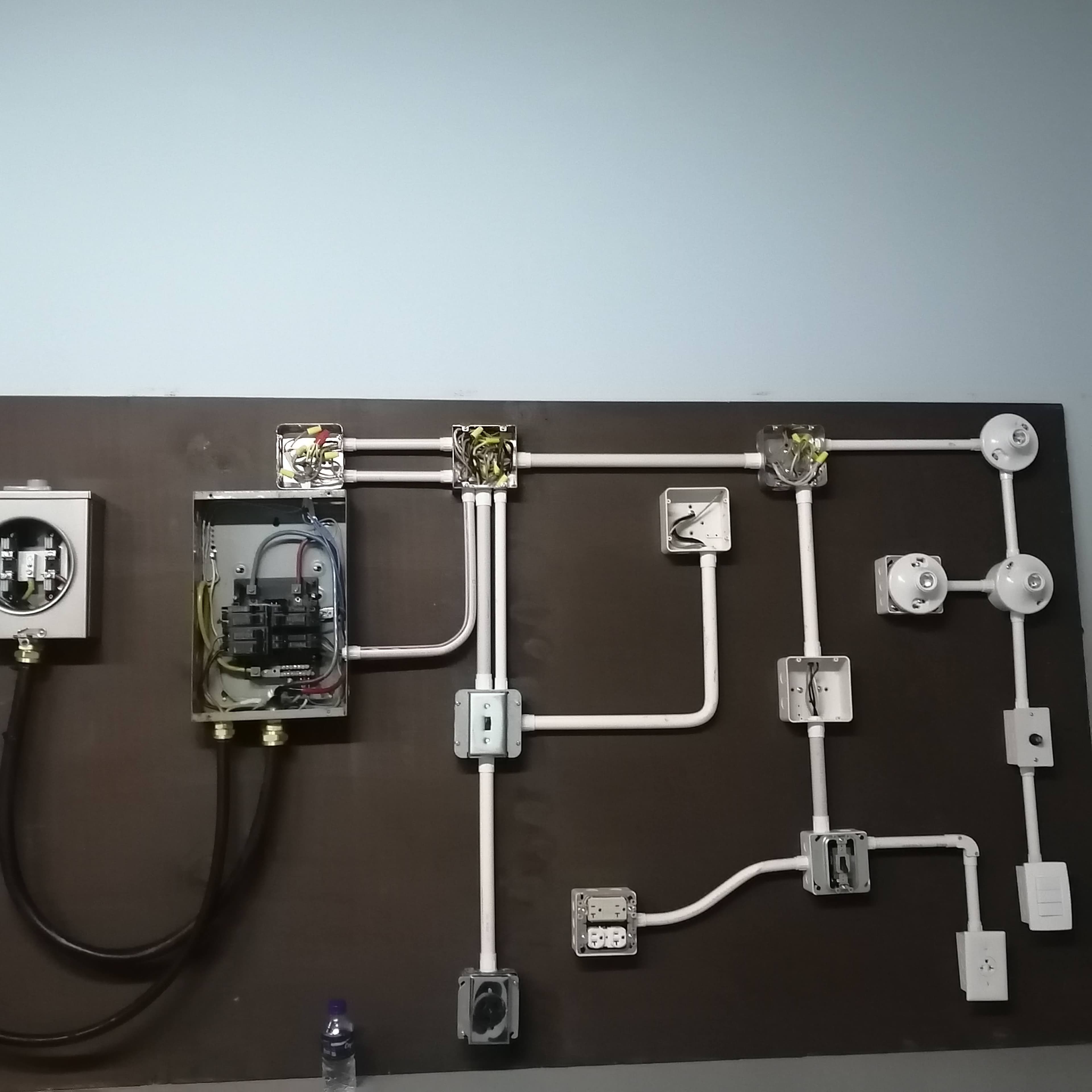 Electrical training board with circuit breaker panel, white conduits, junction boxes, and electrical fixtures.