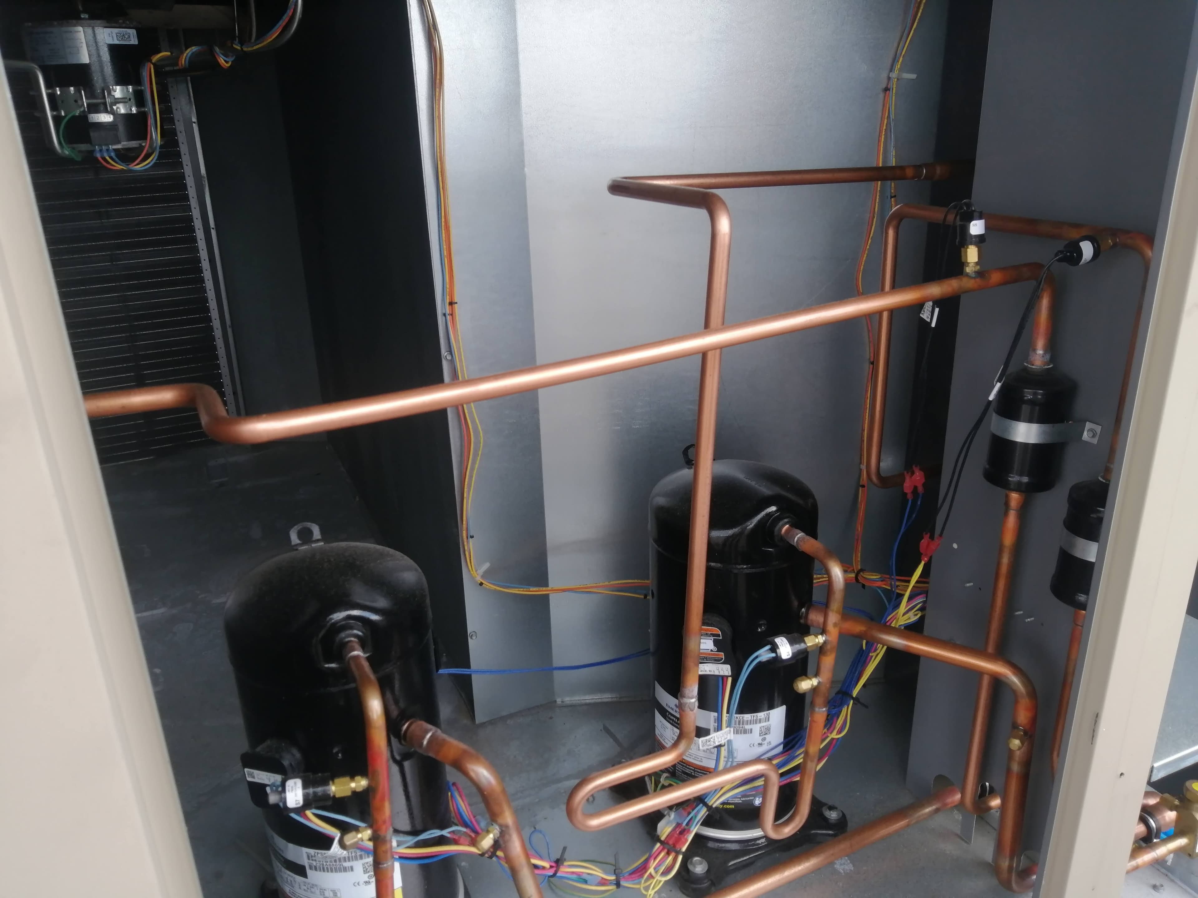 Two black compressors, copper piping, and colorful electrical wiring inside an HVAC unit cabinet.