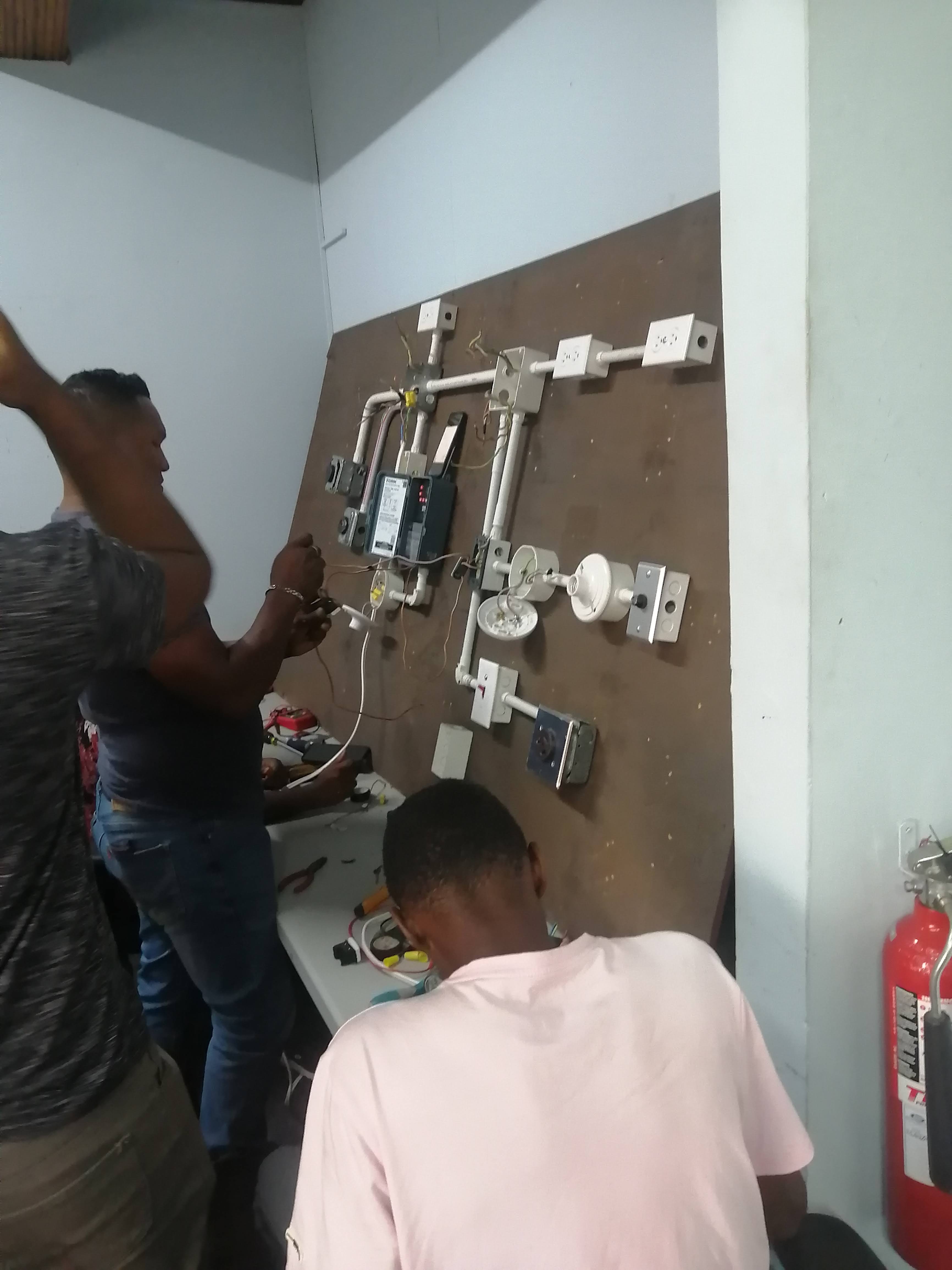 Two men working on a complex electrical wiring board with various outlets and conduits.