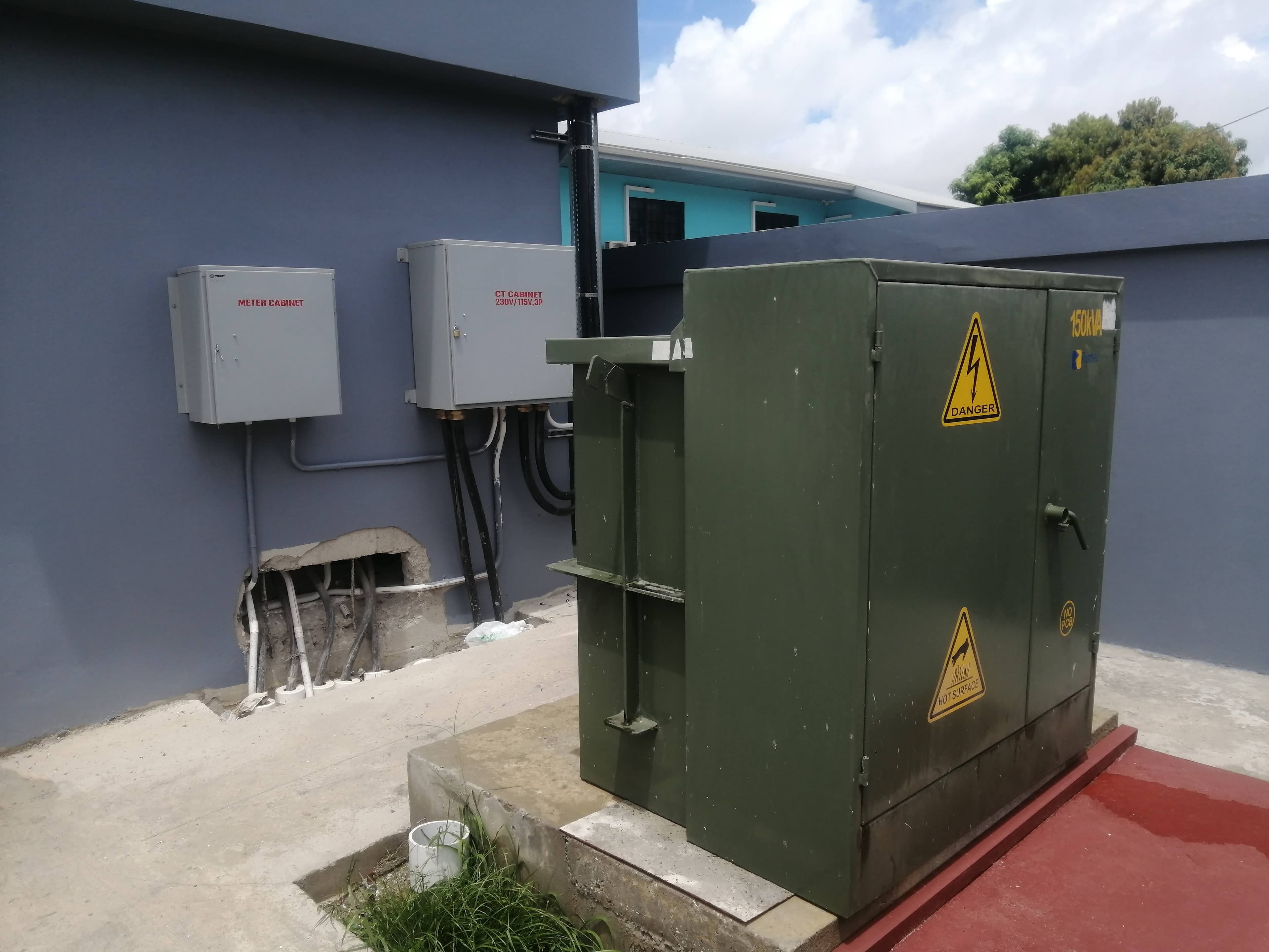 Green electrical transformer box next to grey meter cabinets on a blue exterior wall.