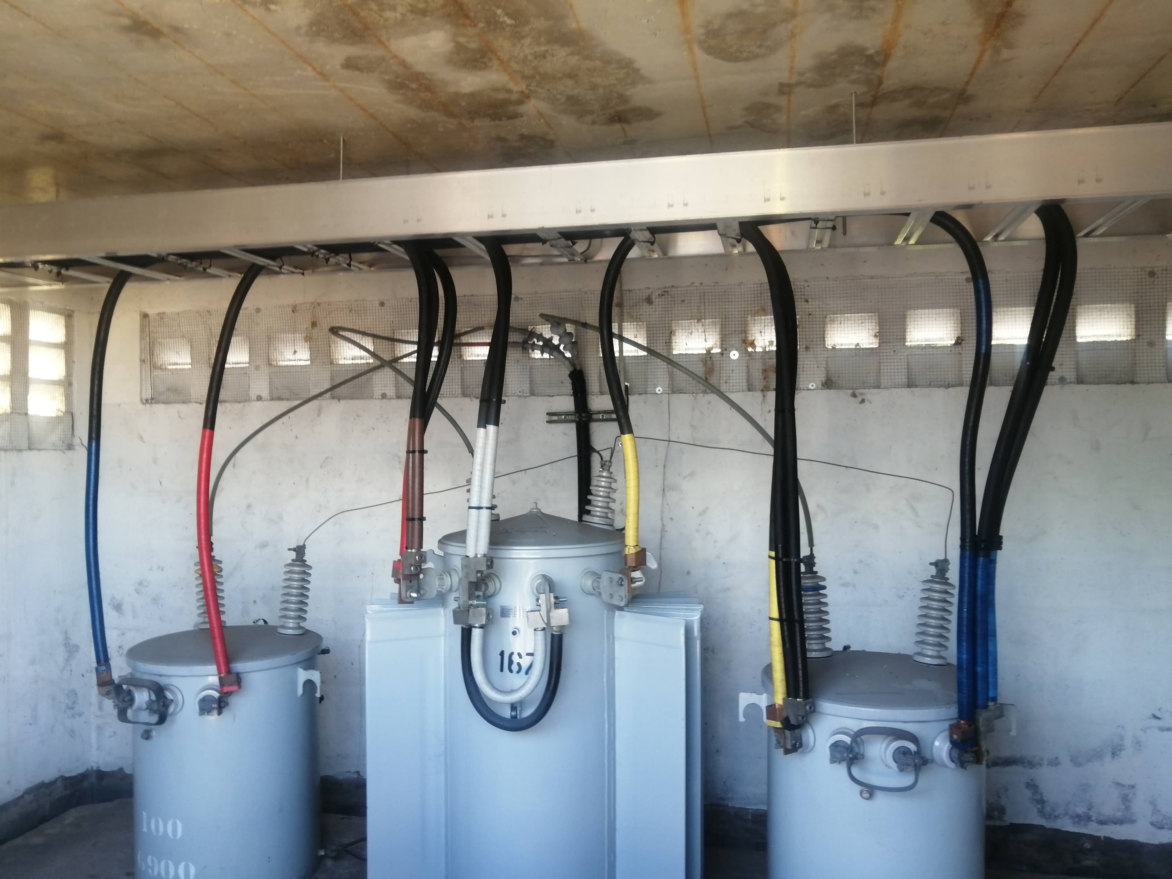 Three large gray electrical transformers with thick cables connected to an overhead metal tray.
