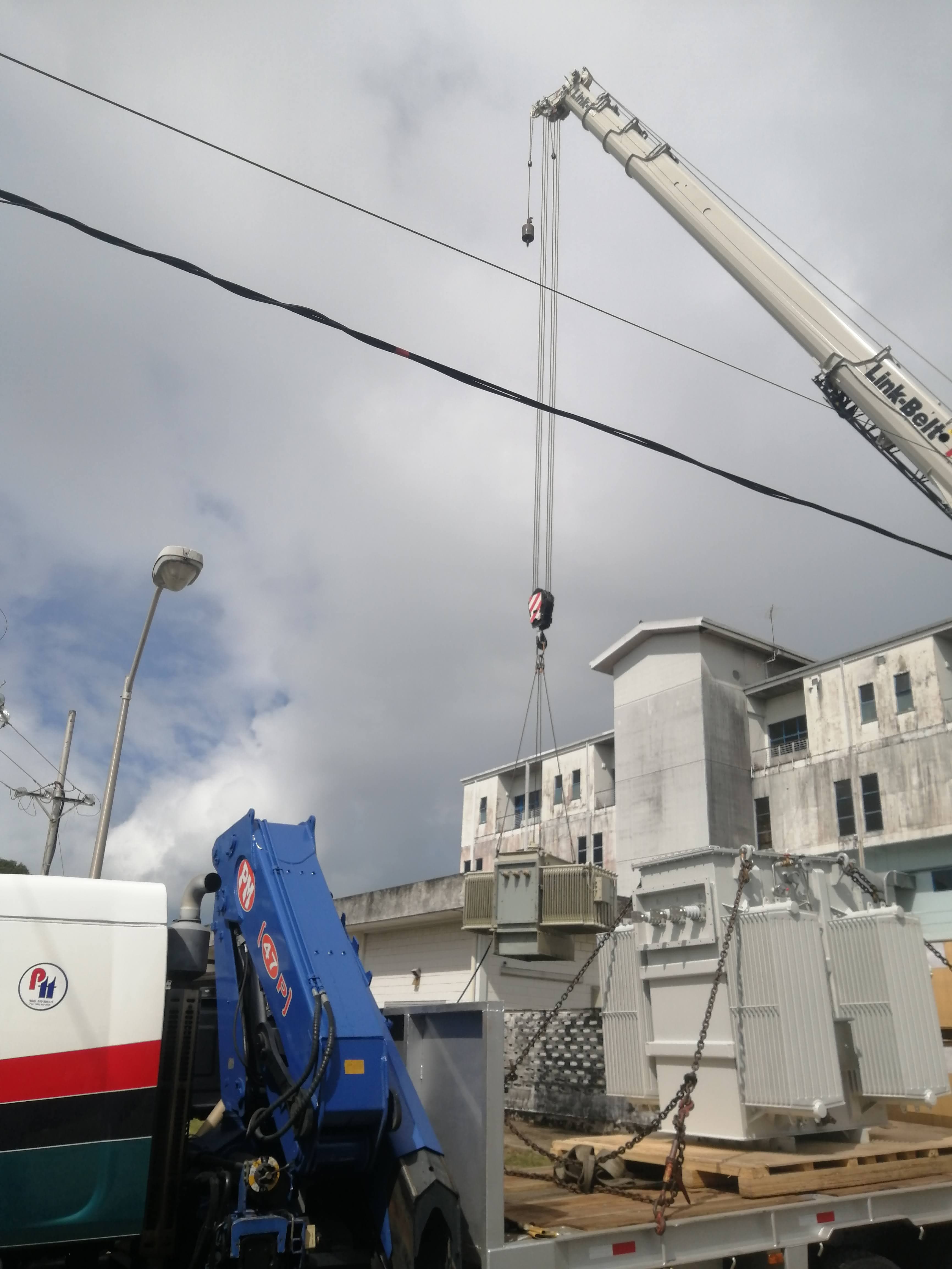 White crane lowering a large grey electrical transformer onto a flatbed truck near buildings.
