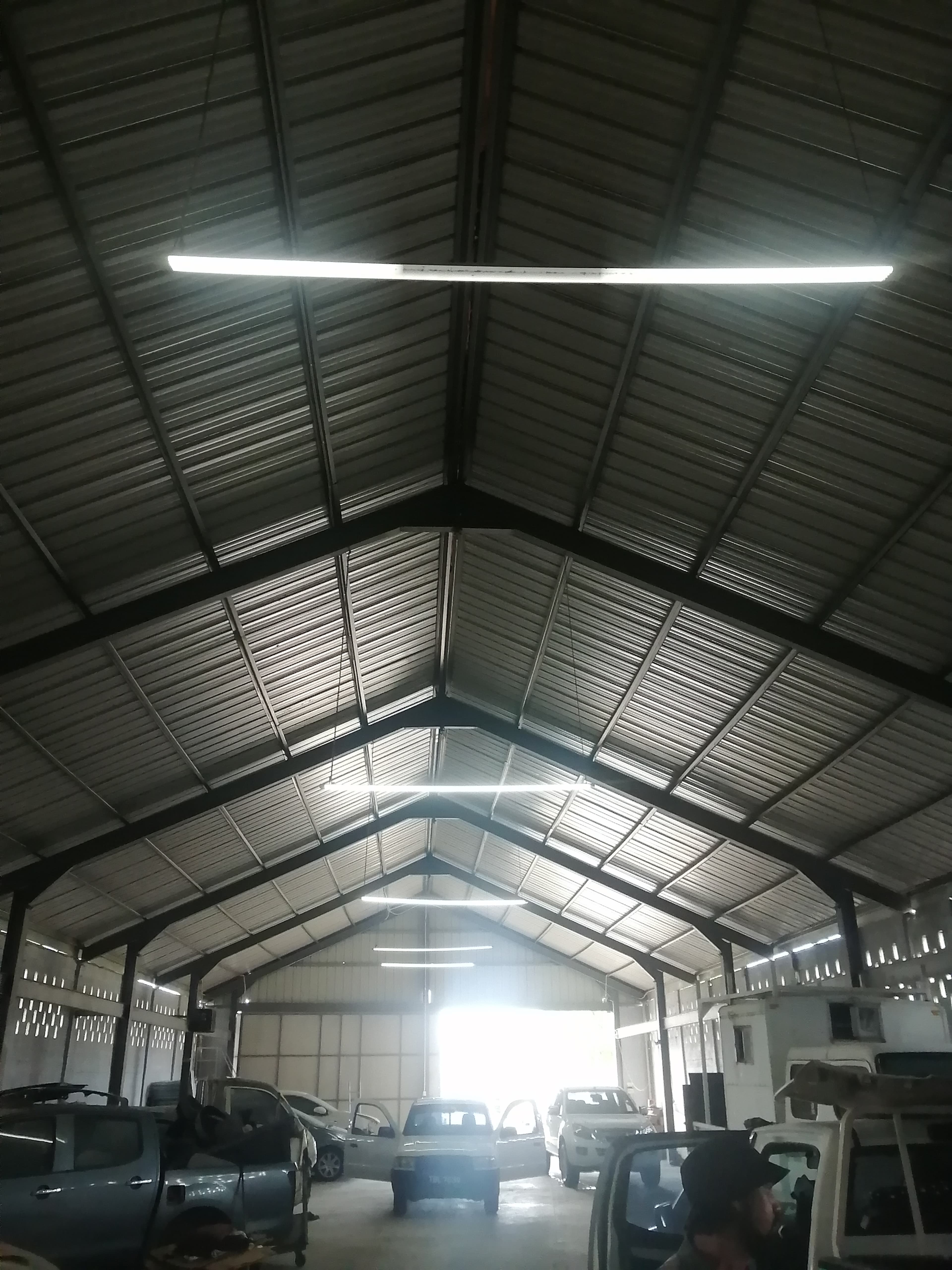 Interior of a corrugated metal warehouse with vehicles parked under several long fluorescent lights.