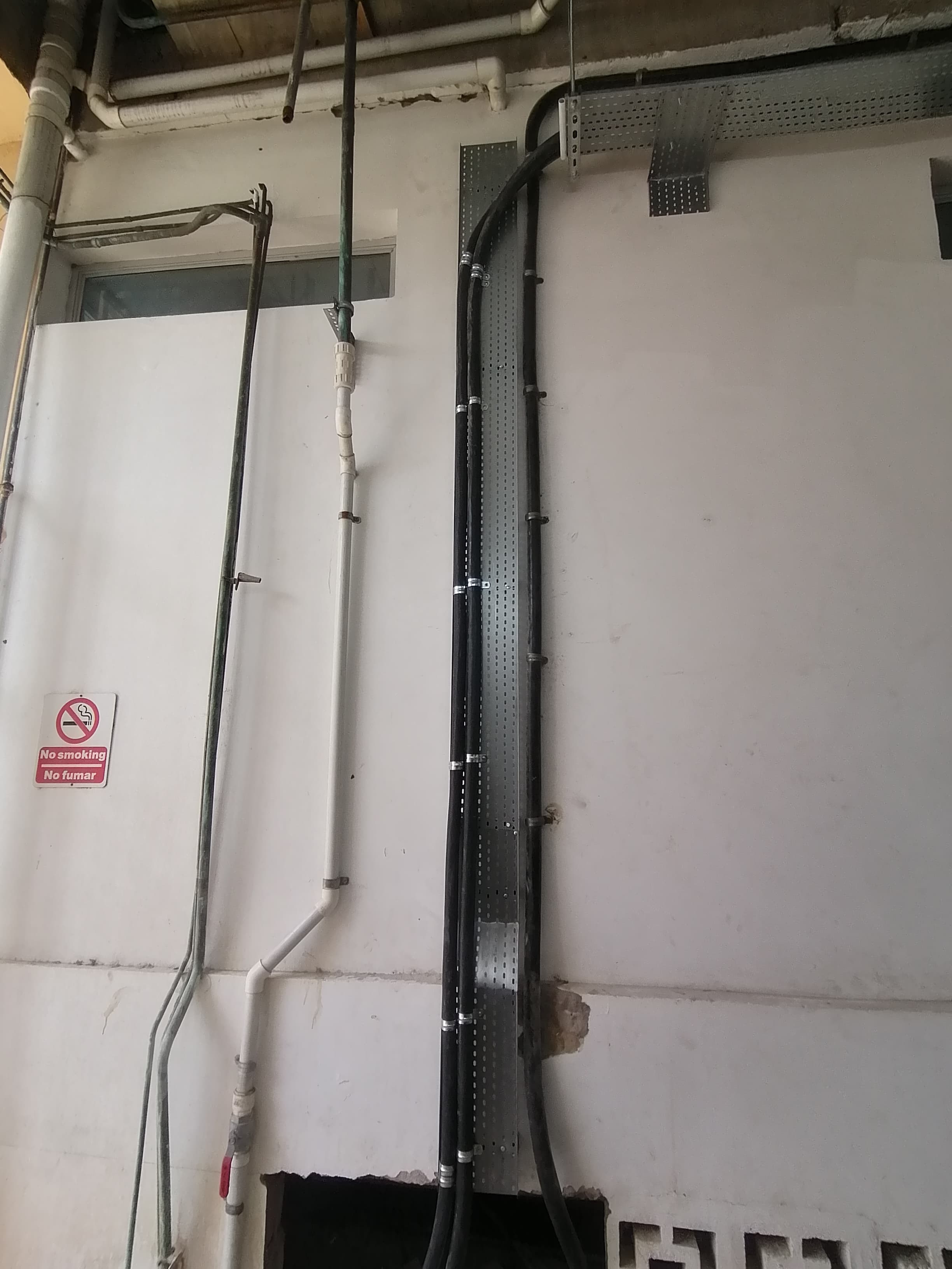 Black cables and industrial pipes on a white wall with a bilingual no smoking sign.